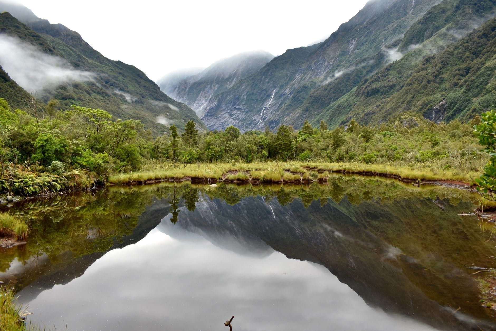 Ein ruhiger Bergsee spiegelt die umliegenden bewaldeten Berge und das neblige Tal des Franz Josef Glacier.