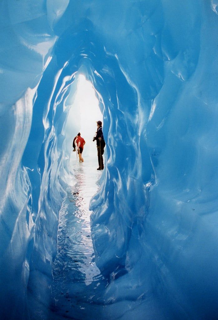 Hikers stand inside a narrow, deep blue glacier ice cave in New Zealand.