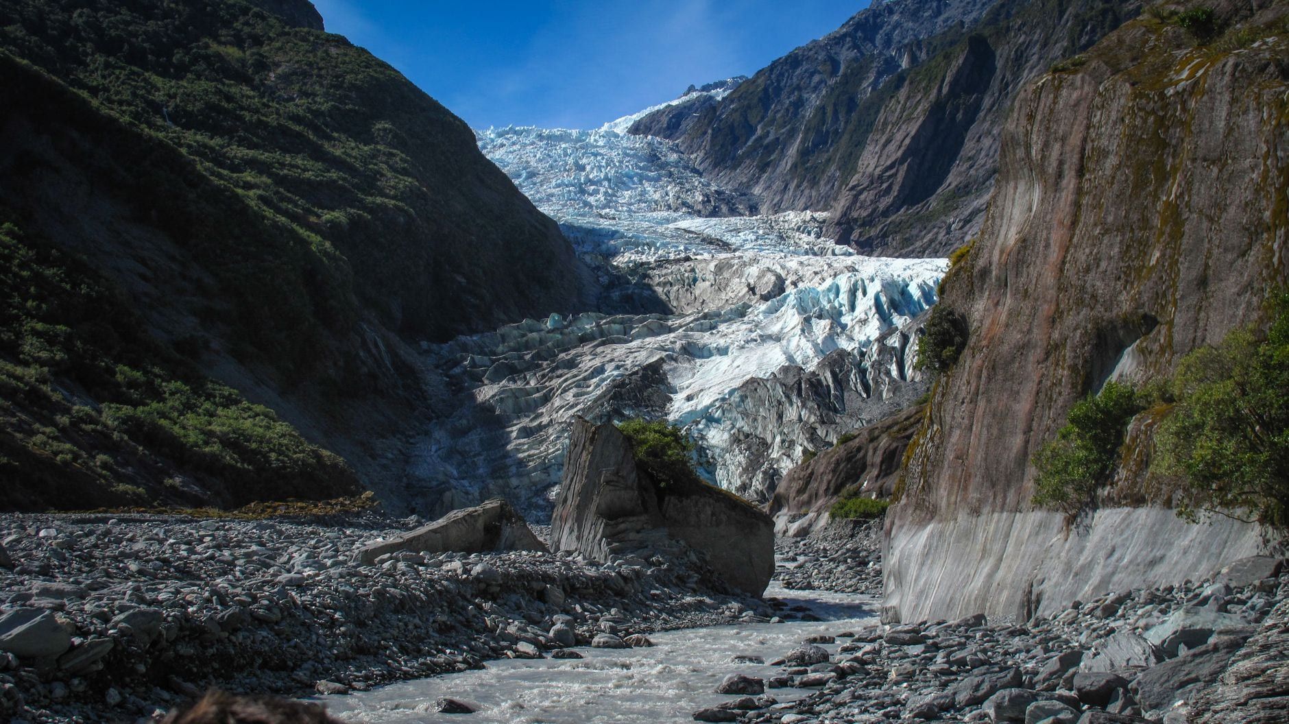 Gletscherfluss, der durch ein felsiges Tal zum Eis des Franz Josef Gletschers fließt, gerahmt von steilen Berghängen.