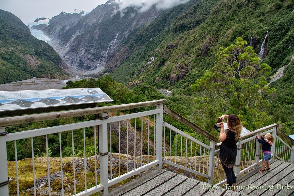 Visitors stand on the Sentinel Rock viewing deck beside an interpretive panel overlooking Franz Josef Glacier.