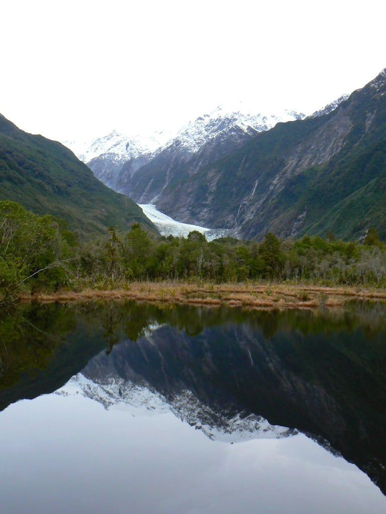 Franz Josef Glacier and snow-dusted mountains reflected in the calm waters of Peters Pool.