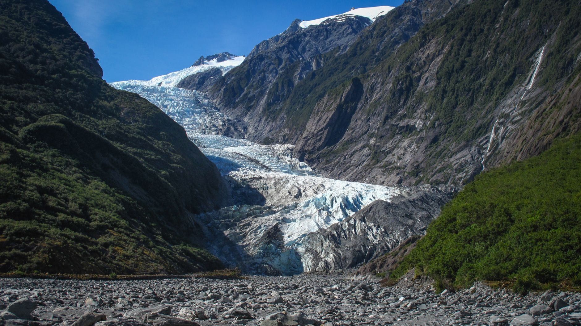 Der Franz Josef Glacier fließt durch eine steile Bergschlucht, gerahmt von felsigen Klippen und üppig grünen Hängen unter blauem Himmel.