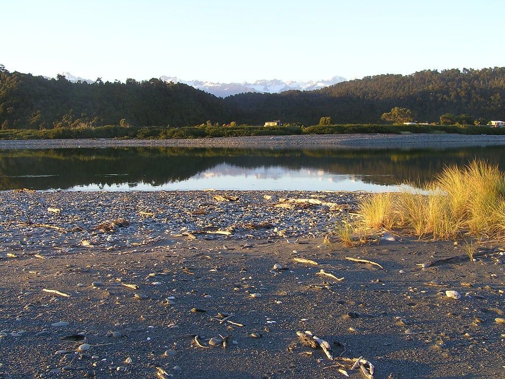 Las aguas tranquilas de la laguna de Okarito reflejan colinas boscosas y los lejanos picos nevados de los Alpes Neozelandeses, cerca del glaciar Franz Josef.