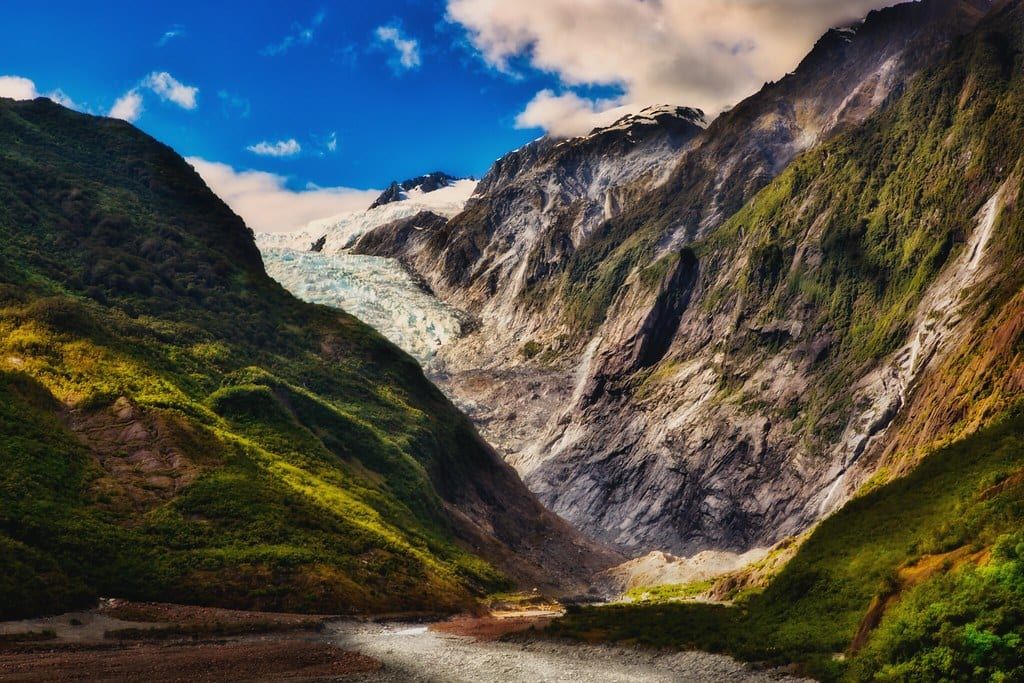 Ein weiter Talblick auf den Franz Josef Glacier, gerahmt von steilen Bergwänden unter blauem, wolkigem Himmel.